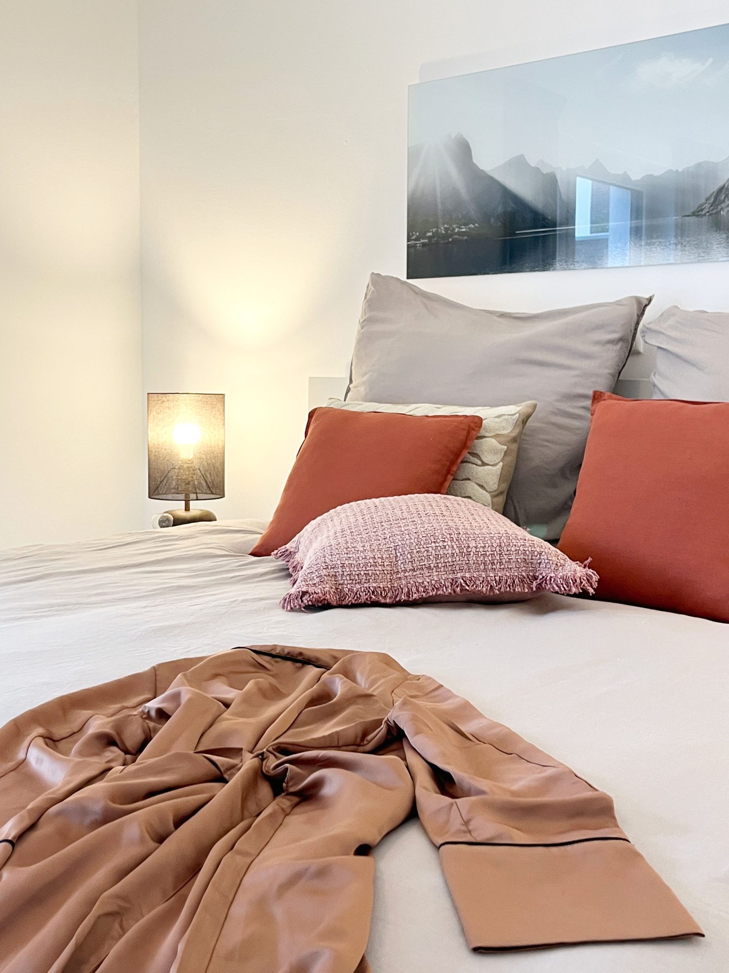 Ferienwohnung_Pamelarohmeyer Auschnitt eines Bettes mit heller Tagesdecke und verschiedenen Kissen. Auf dem Bett liegt ein Satinbademantel