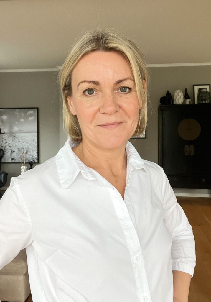 Pamelarohmeyer_diekammerherrin Der Oberkörper einer blonden Frau mit halblangen Haaren in einer weißen Bluse. Die Hände sind auf den Hüften gestützt.